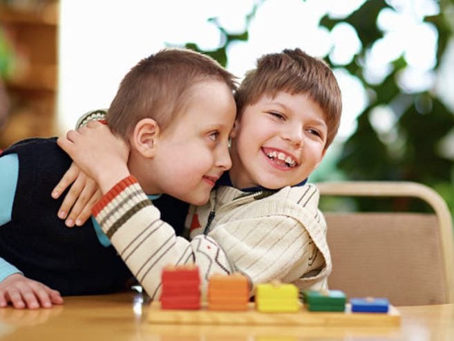 Two young boys sit at a table, smiling and hugging each other warmly. Brightly colored wooden blocks are in front of them. The background is blurred with green plants visible.