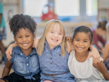 Three young girls sit closely together in a classroom, smiling with their arms around each other. They appear happy and friendly, representing diversity and inclusion.