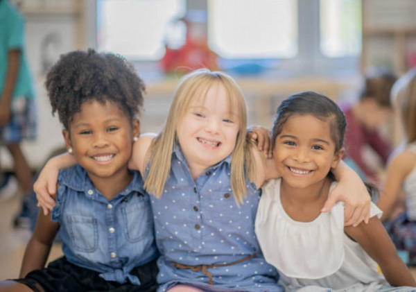 Three young girls sit closely together in a classroom, smiling with their arms around each other. They appear happy and friendly, representing diversity and inclusion.