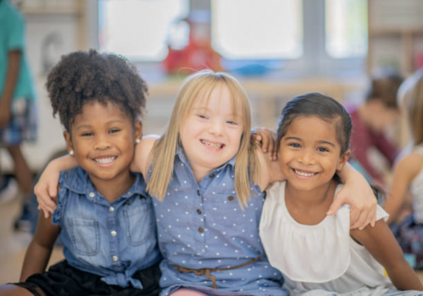 Three young girls sit closely together in a classroom, smiling with their arms around each other. They appear happy and friendly, representing diversity and inclusion.
