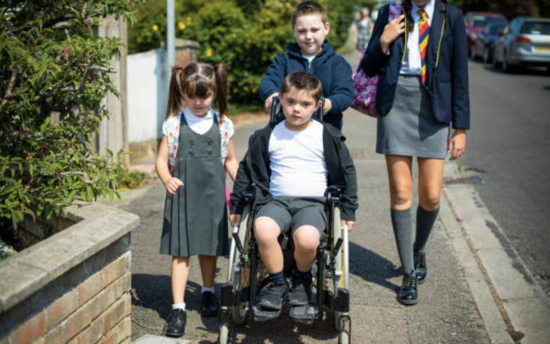 Four children walk along a suburban sidewalk, one child in a wheelchair pushed by another, with two others walking beside them. They are wearing school uniforms and carry backpacks.