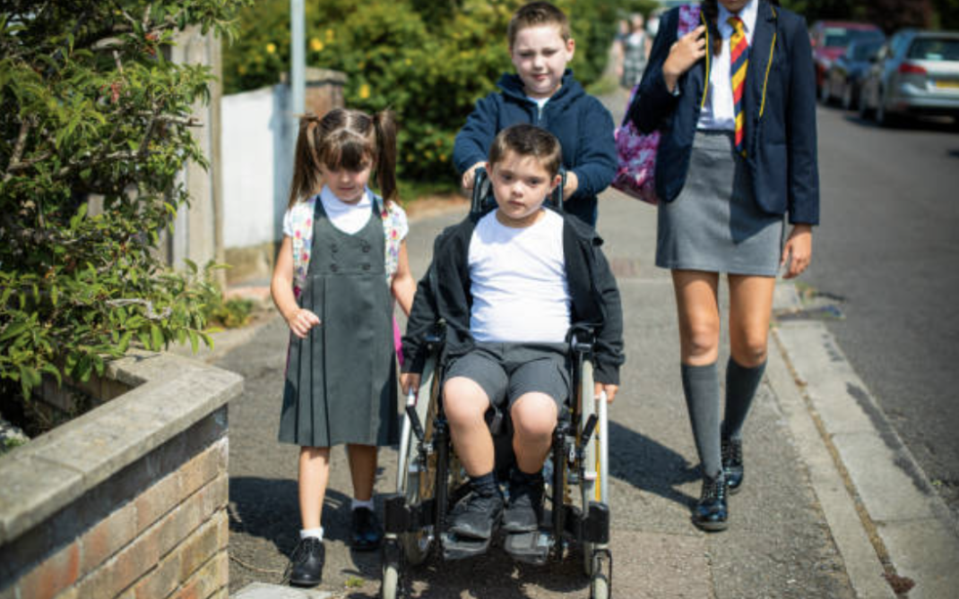 Four children walk along a suburban sidewalk, one child in a wheelchair pushed by another, with two others walking beside them. They are wearing school uniforms and carry backpacks.