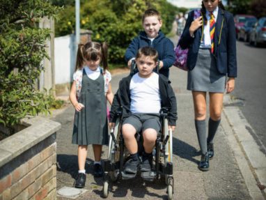 Four children walk along a suburban sidewalk, one child in a wheelchair pushed by another, with two others walking beside them. They are wearing school uniforms and carry backpacks.