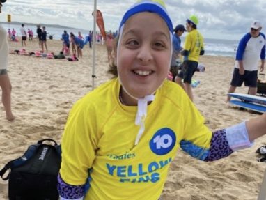 A smiling child wearing a yellow Yellow Fins rash guard, swim cap, and patterned sleeves stands on a sandy beach with other people and surfboards in the background.