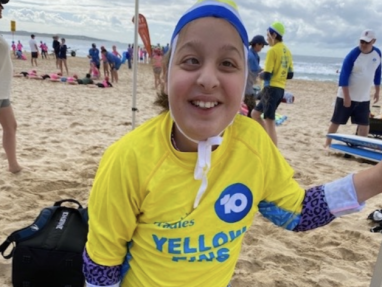 A smiling child wearing a yellow Yellow Fins rash guard, swim cap, and patterned sleeves stands on a sandy beach with other people and surfboards in the background.