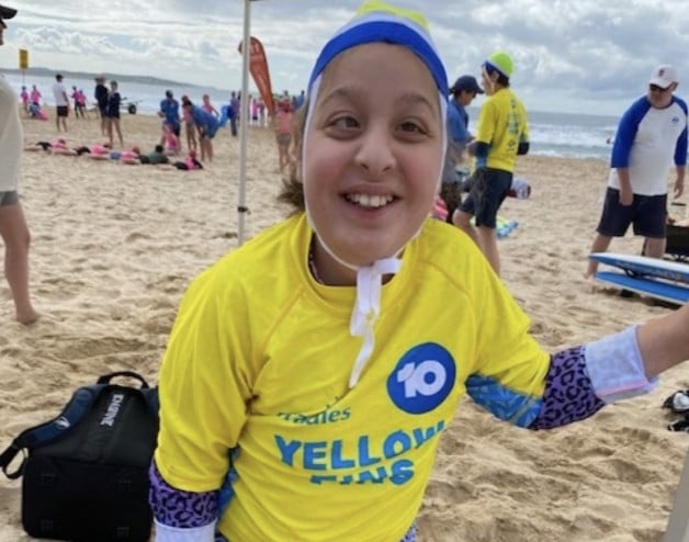 A smiling child wearing a yellow Yellow Fins rash guard, swim cap, and patterned sleeves stands on a sandy beach with other people and surfboards in the background.