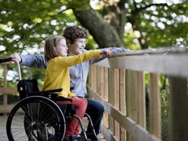 A young girl in a wheelchair and a boy stand together on a wooden boardwalk, both smiling and pointing at something in the distance, surrounded by green trees.