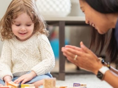 A young child in a white sweater sits on the floor playing with colorful alphabet blocks while an adult nearby smiles and claps, encouraging the child.