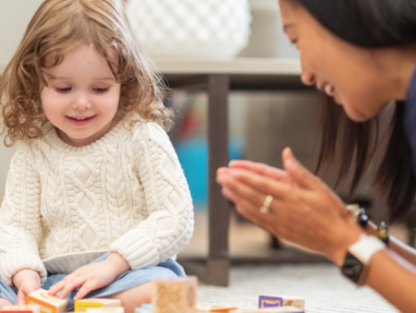 A young child in a white sweater sits on the floor playing with colorful alphabet blocks while an adult nearby smiles and claps, encouraging the child.