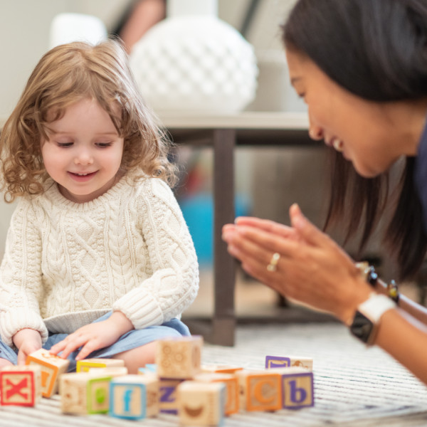 A young child in a white sweater sits on the floor playing with colorful alphabet blocks while an adult nearby smiles and claps, encouraging the child.