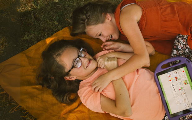 Two girls lying on an orange blanket outdoors, smiling and playing together. One girl wears glasses and holds a purple speech-generating device with icons on its screen.