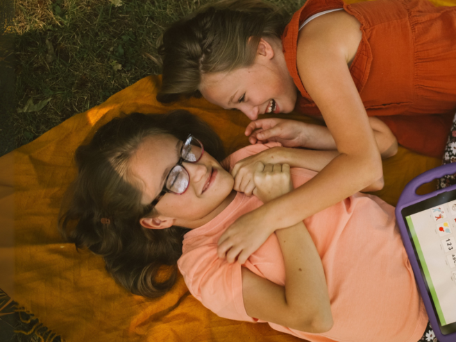 Two girls lying on an orange blanket outdoors, smiling and playing together. One girl wears glasses and holds a purple speech-generating device with icons on its screen.