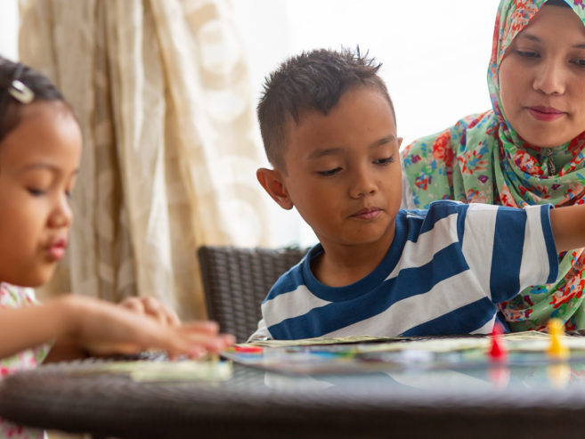 A woman and two young children sit at a round table playing a board game together. The woman wears a patterned hijab, and the children are focused on the game pieces. Sunlight streams through a window behind them.