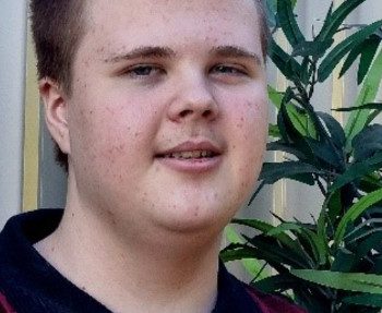 A young person with short brown hair, wearing a maroon and black collared shirt, stands outdoors in front of a metal fence and green leafy plant, smiling softly at the camera.