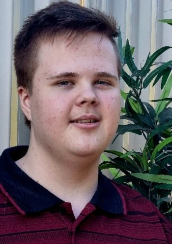 A young person with short brown hair, wearing a maroon and black collared shirt, stands outdoors in front of a metal fence and green leafy plant, smiling softly at the camera.
