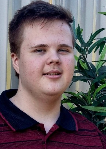 A young person with short brown hair, wearing a maroon and black collared shirt, stands outdoors in front of a metal fence and green leafy plant, smiling softly at the camera.