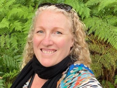 A woman with curly blonde hair, wearing sunglasses on her head, a black scarf, and a colorful top, smiles at the camera while standing in front of lush green ferns in an outdoor setting.