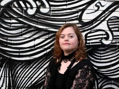 A young woman with Down syndrome, wearing a black lace top, stands in front of a black and white abstract mural with swirling lines and shapes. She looks confidently at the camera.