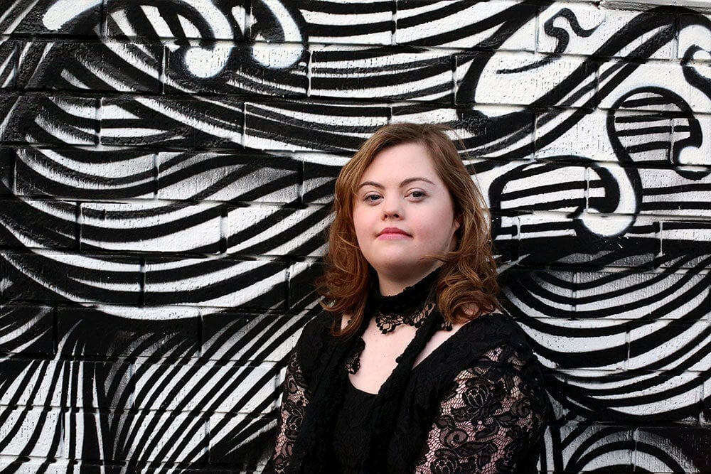 A young woman with Down syndrome, wearing a black lace top, stands in front of a black and white abstract mural with swirling lines and shapes. She looks confidently at the camera.