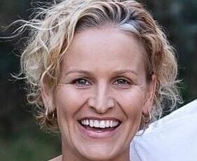 A woman with short, curly blonde hair smiles widely at the camera, standing outdoors with a blurred green background.