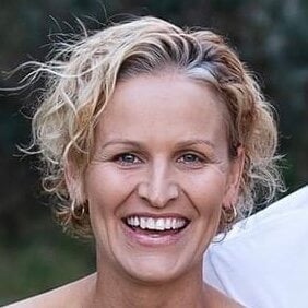 A woman with short, curly blonde hair smiles widely at the camera, standing outdoors with a blurred green background.