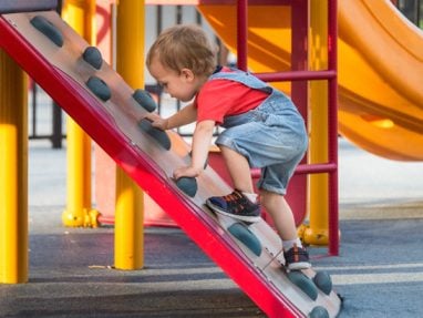 A young child in a red shirt and denim overalls climbs up a small plastic rock wall on a playground, surrounded by colorful play equipment.