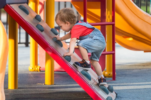 A young child in a red shirt and denim overalls climbs up a small plastic rock wall on a playground, surrounded by colorful play equipment.