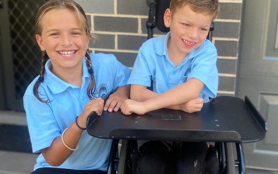 Two smiling children in light blue school uniforms, one sitting in a wheelchair and the other standing beside them, posing together in front of a brick wall and door.
