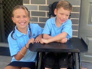 Two smiling children in light blue school uniforms, one sitting in a wheelchair and the other standing beside them, posing together in front of a brick wall and door.