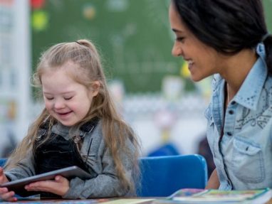 A young girl with Down syndrome smiles as she looks at a tablet, while a woman sitting beside her also smiles. They are seated at a table in a colorful classroom.