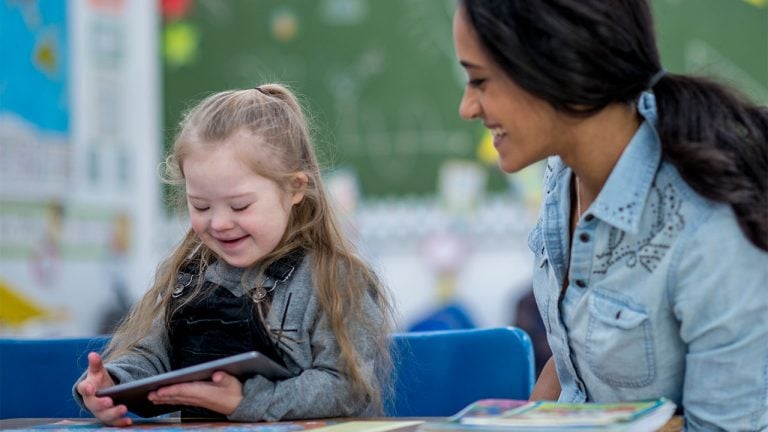 A young girl with Down syndrome smiles as she looks at a tablet, while a woman sitting beside her also smiles. They are seated at a table in a colorful classroom.