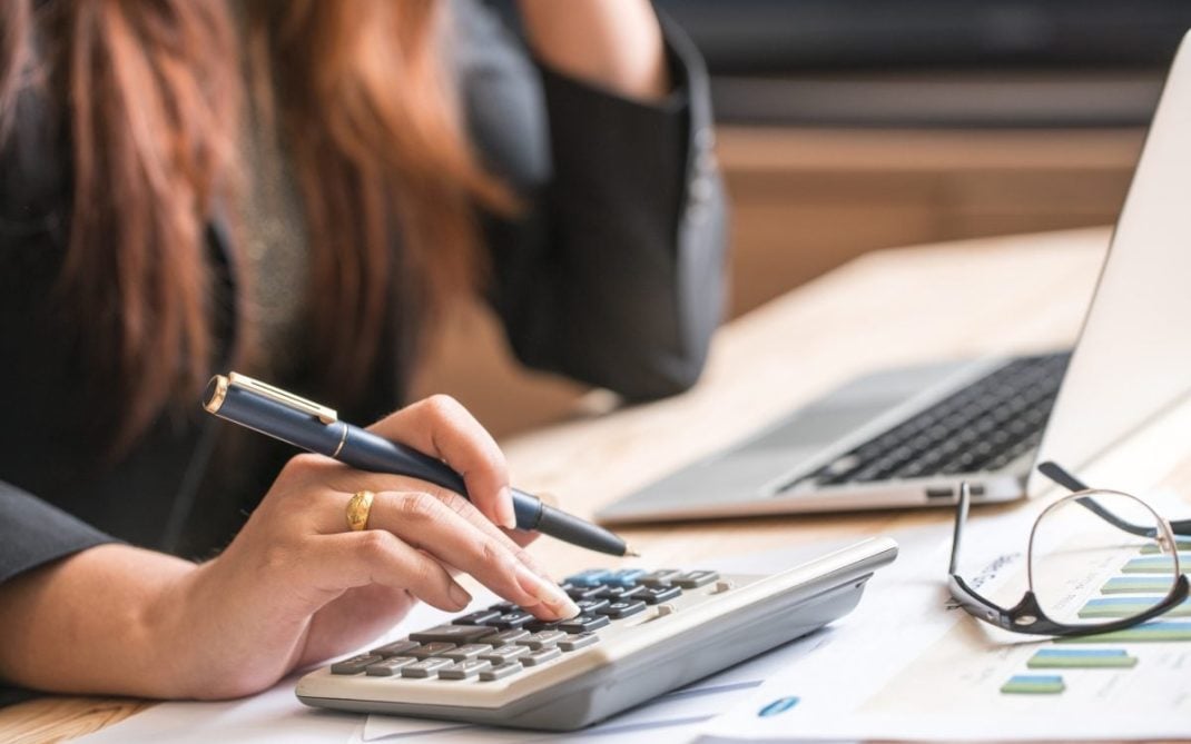 A person using a calculator with one hand while holding a pen in the other, seated at a desk with documents, eyeglasses, and a laptop.
