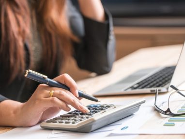A person using a calculator with one hand while holding a pen in the other, seated at a desk with documents, eyeglasses, and a laptop.