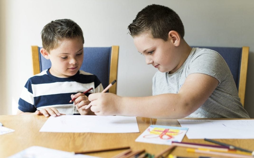 Two boys sit at a wooden table drawing with colored pencils on white paper. One boy in a striped shirt watches while the other, in a gray shirt, draws. Art supplies and finished drawings are scattered on the table.