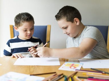 Two boys sit at a wooden table drawing with colored pencils on white paper. One boy in a striped shirt watches while the other, in a gray shirt, draws. Art supplies and finished drawings are scattered on the table.