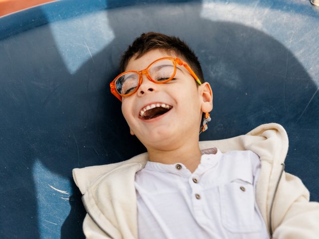A young boy wearing orange glasses and a white shirt lies on a blue surface, smiling and laughing with his eyes closed, enjoying a playful moment.