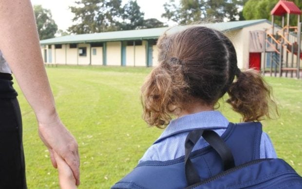 A young child with pigtails and a backpack holds an adult’s hand while walking toward a school building, with a playground visible to the right and green grass surrounding them.