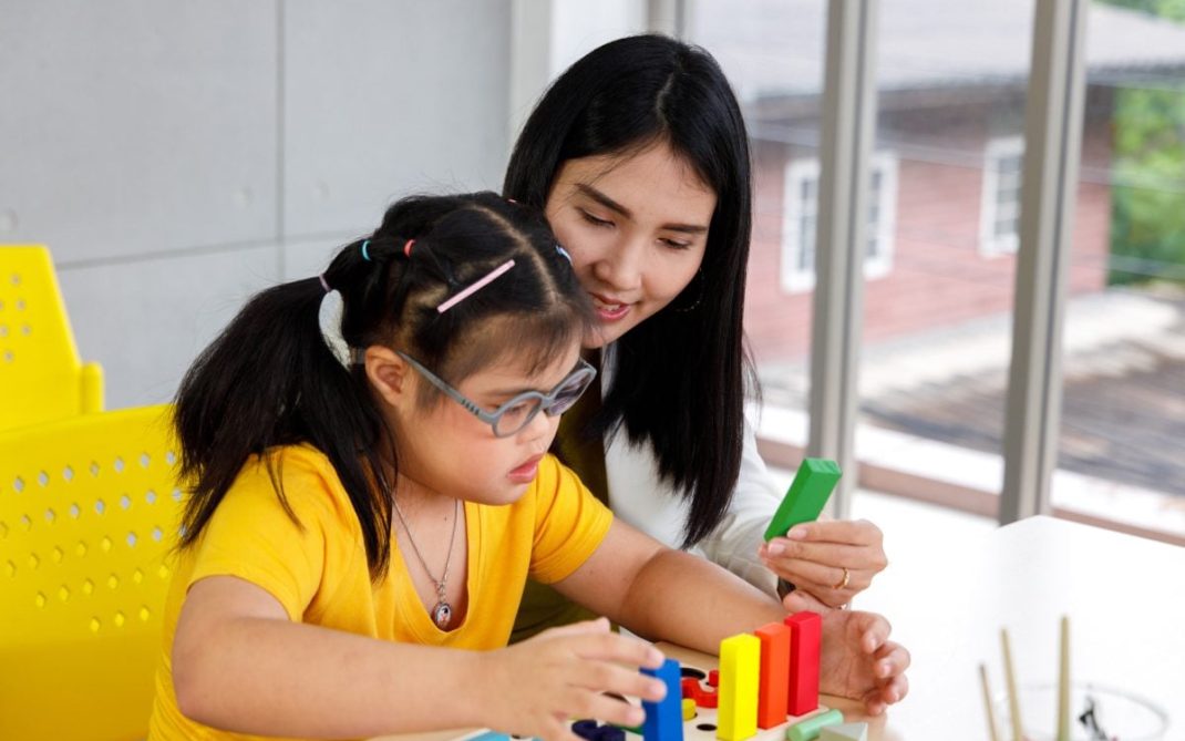 A young girl wearing glasses arranges colorful blocks while a woman beside her watches and smiles, both seated at a table in a bright room with yellow chairs and large windows.