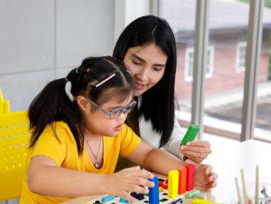 A young girl wearing glasses arranges colorful blocks while a woman beside her watches and smiles, both seated at a table in a bright room with yellow chairs and large windows.