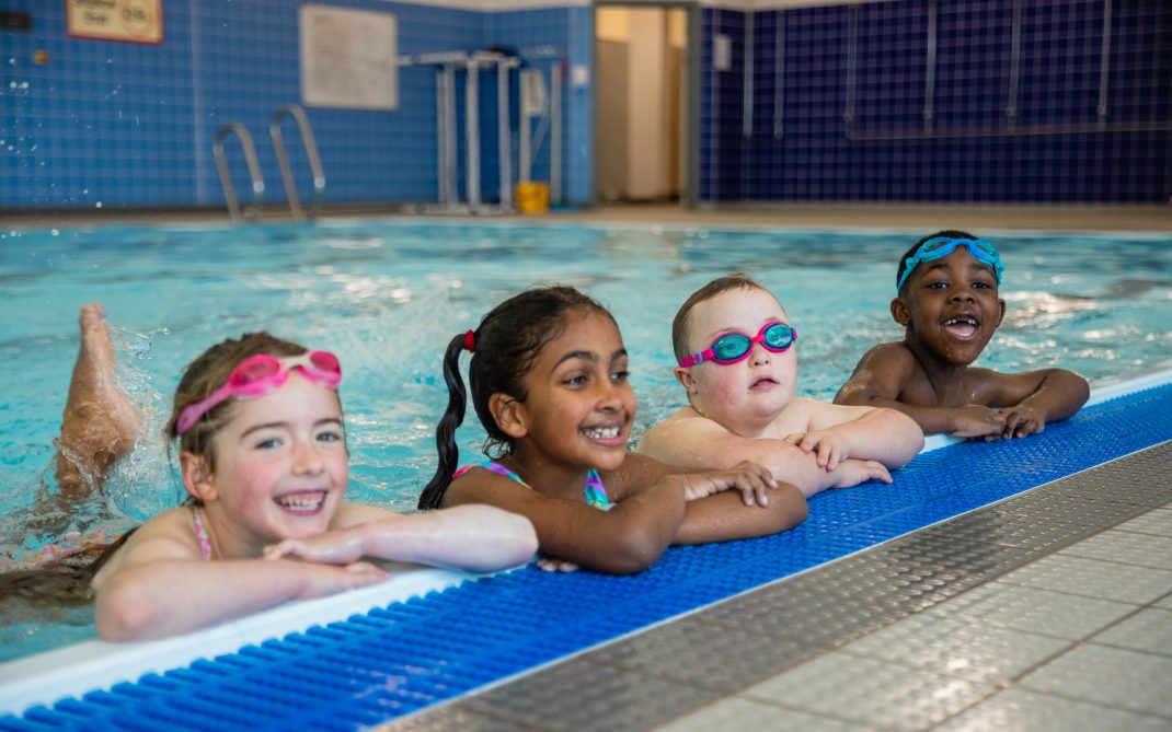 Four young children in swimsuits and goggles smile and rest their arms on the edge of an indoor swimming pool, with blue tiles and pool equipment visible in the background.