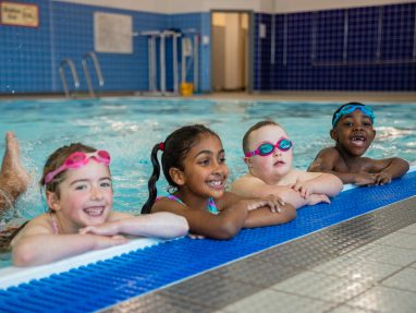 Four young children in swimsuits and goggles smile and rest their arms on the edge of an indoor swimming pool, with blue tiles and pool equipment visible in the background.