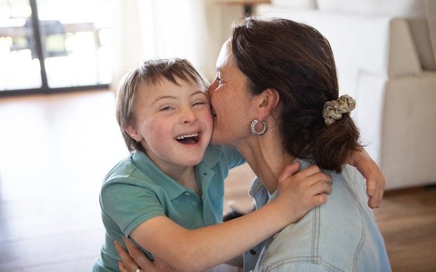 A joyful young boy with Down syndrome smiles as a woman hugs and kisses him on the cheek in a bright living room.