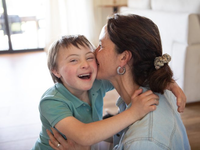 A joyful young boy with Down syndrome smiles as a woman hugs and kisses him on the cheek in a bright living room.