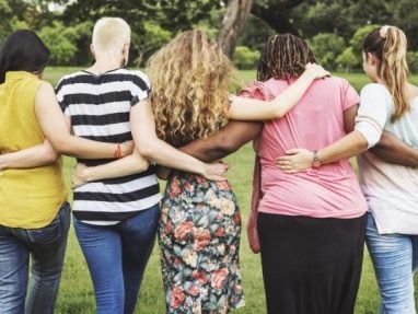 Five women stand outdoors with their backs to the camera, arms around each other’s shoulders, showing friendship and support. They are in a grassy area with trees in the background.