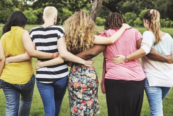 Five women stand outdoors with their backs to the camera, arms around each other’s shoulders, showing friendship and support. They are in a grassy area with trees in the background.