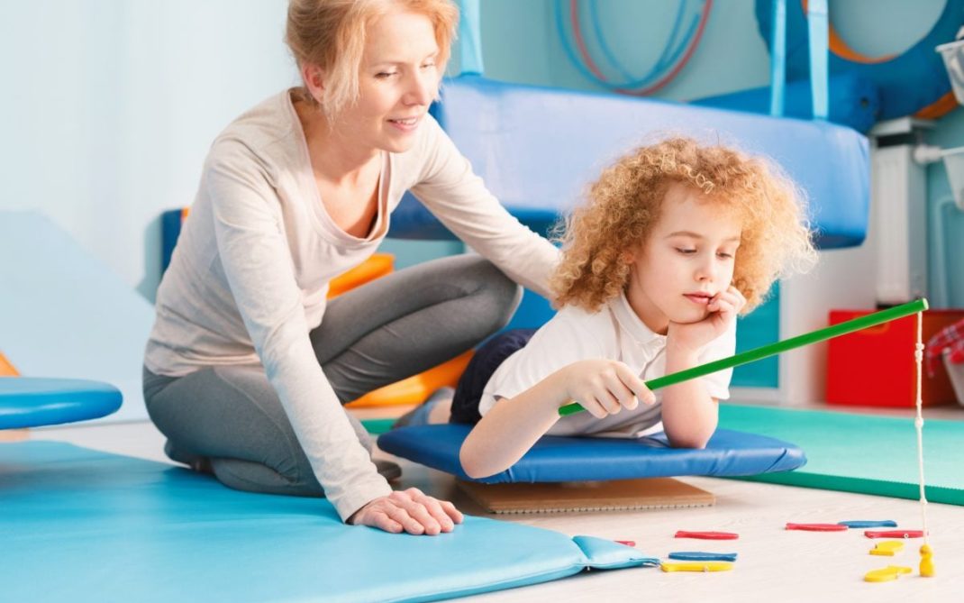 A woman assists a young child lying on a blue mat, holding a green stick with string and colorful objects on the floor, in a brightly lit therapy or playroom setting.