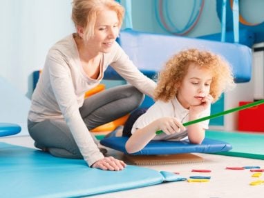 A woman assists a young child lying on a blue mat, holding a green stick with string and colorful objects on the floor, in a brightly lit therapy or playroom setting.