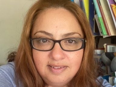 A woman with light brown hair and glasses smiles at the camera, wearing a grey top and a heart-shaped necklace, with colorful folders and office supplies on a shelf behind her.