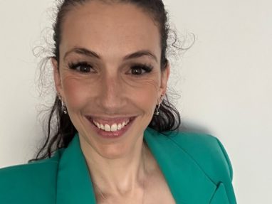A woman with long brown hair smiles at the camera. She is wearing a teal blazer, a light-colored top, hoop earrings, and a silver necklace, standing against a plain white background.
