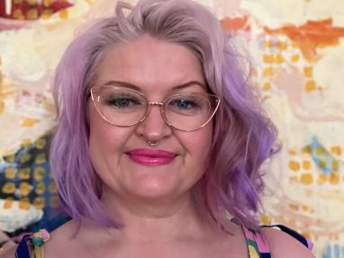 A person with pastel purple hair, cat-eye glasses, and a nose ring smiles in front of a colorful abstract background. They wear a striped top with thin straps and have a visible tattoo on their right arm.